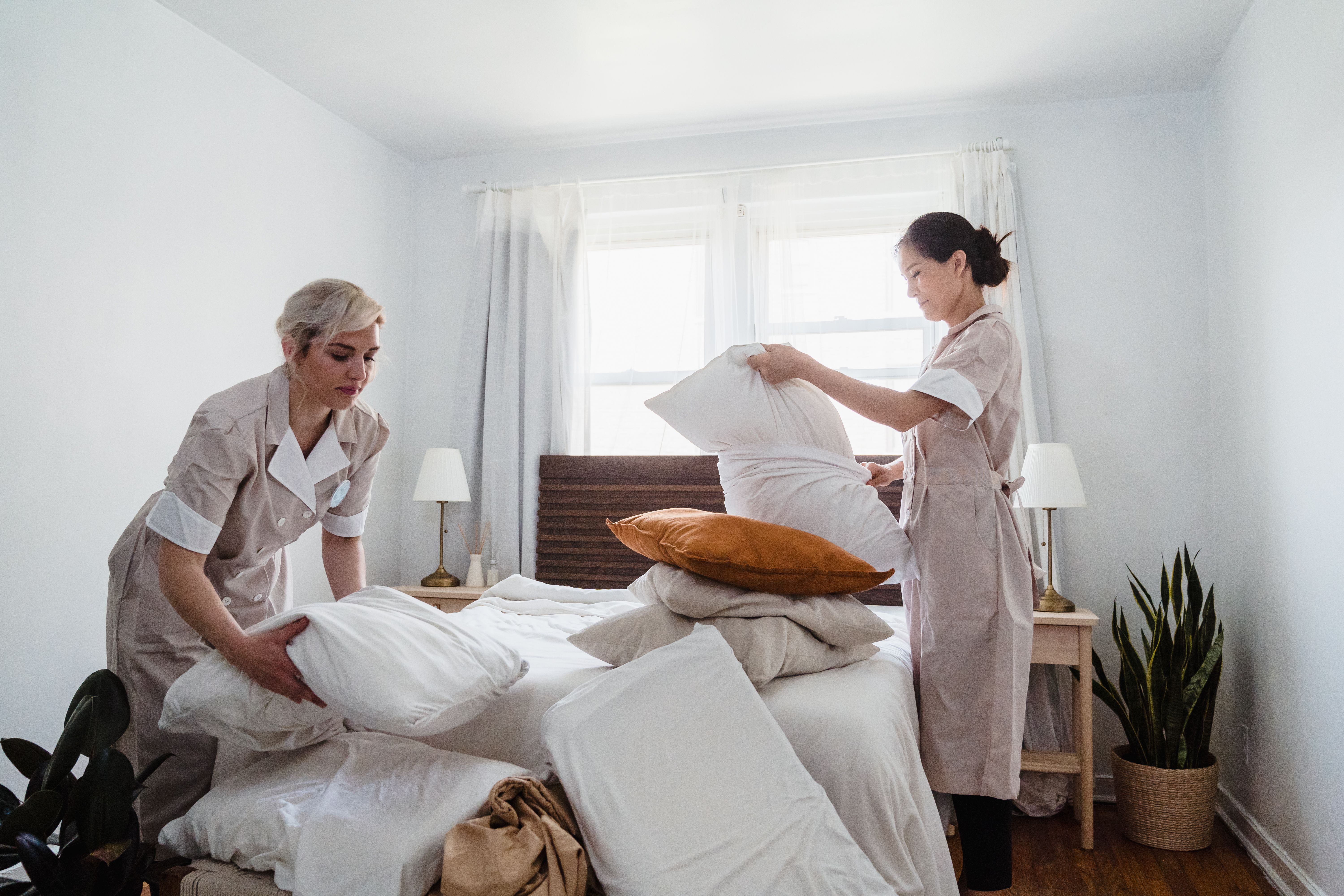 Twee hotelmedewerkers maken een bed op in een lichte hotelkamer.